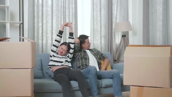 Asian Couple Carrying Cardboard Boxes With Stuff Into A New House Before Sitting On A Sofa To Rest