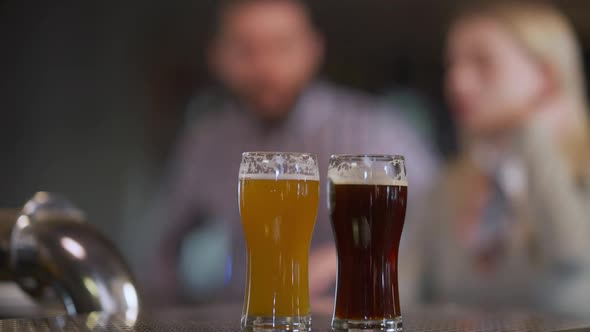 Beer Glasses with Pale and Dark Lager on Bar Counter and Blurred Caucasian Couple Entering Sitting