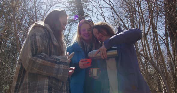 three girls laugh and drink hot tea from a thermos in the forest