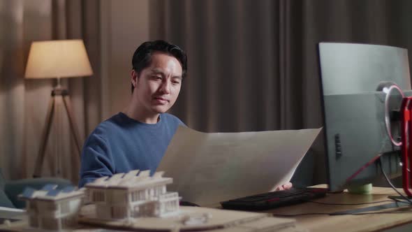 Asian Male Engineer With House Model Opening And Looking At The Blueprint While Working On A Desktop