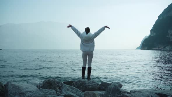 Woman Tourist Raises Hands Standing on Rocks Near Lake Garda