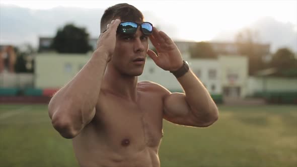 Muscular Man with a Naked Torso After Training Stands in the City Stadium and Puts on Sunglasses