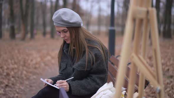 Charming Talented Young Woman Sitting on Bench Drawing Sketch and Looking at Camera Smiling