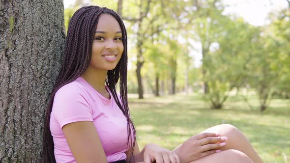 A Young Black Woman Smiles at the Camera As She Sits Under a Tree in a Park on a Sunny Day