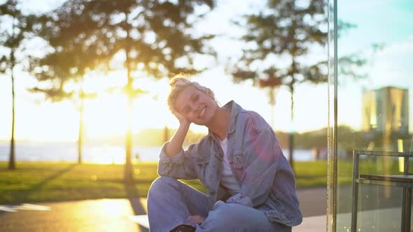 Charming Young Blond Female Student in Denim Sitting in Park Near Riverbank As Sun Shining Behind