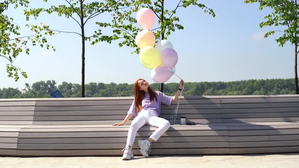 Happy Redhead Young Woman Sitting on Bench with Large Fountain of Colorful Balloons Outside.