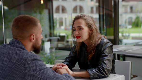 a Man with a Beard and a Blonde with Bright Lipstick Look at Each Other and Hold Hands at a Cafe