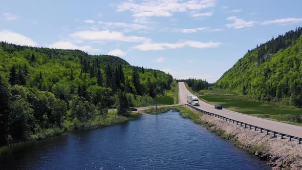 A white semi truck drives down a highway surrounded by forests and mountains next to a lake during t