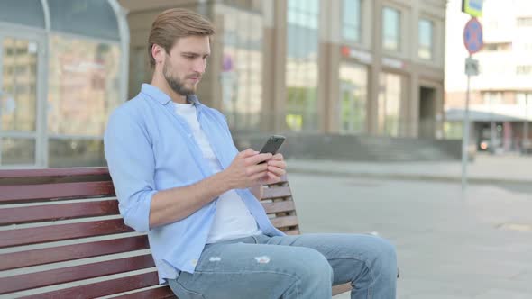 Man Browsing Internet on Smartphone While Sitting Outdoor on Bench