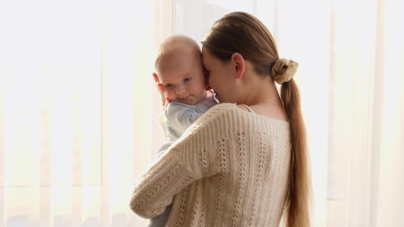 Beautiful Woman Embracing and Kissing Her Little Baby Son Next to Big Window on Sunset