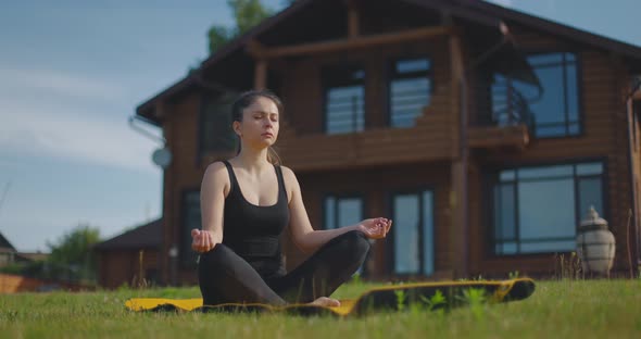 Young Athletic Girl Meditates Sitting on Rug on Lawn Against Background of Country House