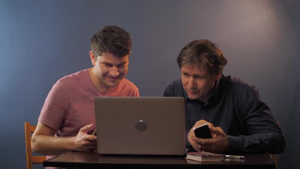 Happy Young and Senior Men with Phones and Laptop at Table