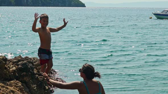 Caucasian kid waves at camera, and jumps into the sea from cape, while his mother watching. Slow mot