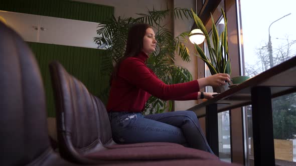 Woman Sits in a Cafeteria Drinking Coffee Near a Large Window to the City