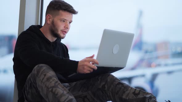 Man Sitting at Airport and Uses Laptop for Work, Blurred Airplane on Background