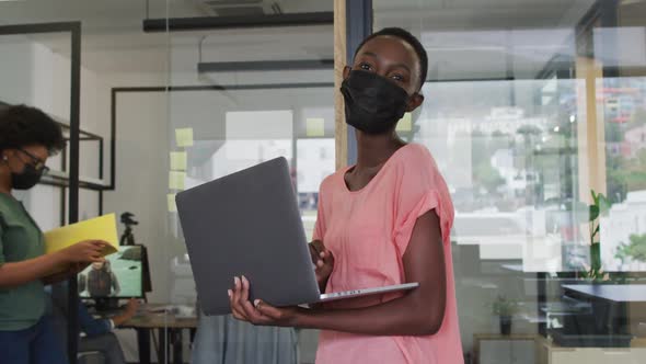 Happy african american businesswoman wearing face mask using laptop, looking to camera in office