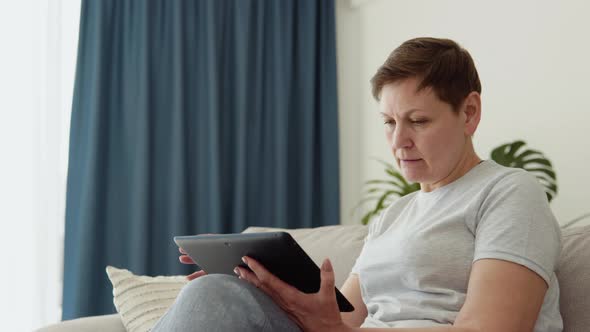 Smiling Elderly Mature Woman Resting on Sofa Using Digital Tablet Alone at Home