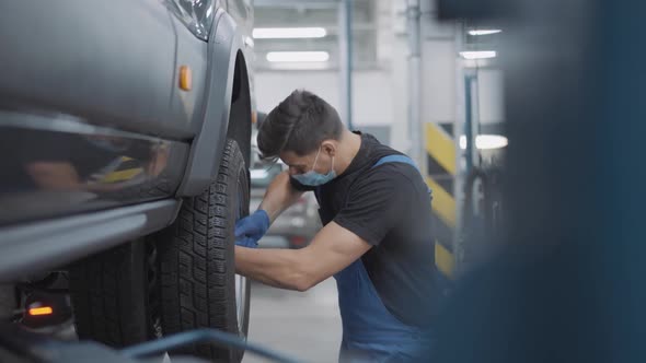 Side View of Auto Mechanic in Covid19 Face Mask Screwing Bolts on Car Wheel