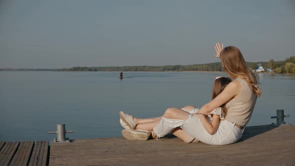 Woman with Daughter Sit on a Pier Waving to Dad Surfing on a Lake