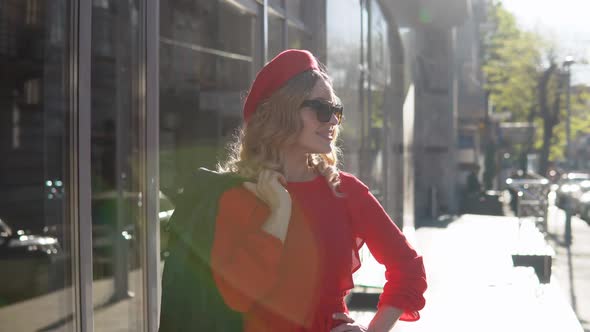 Stylish Young Blonde Woman in a Red Dress and Black Braid Stands on a Cozy Street in the Morning Sun