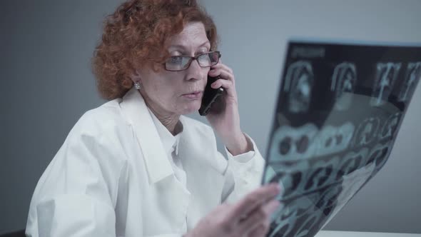 Elderly Caucasian Woman Doctor Analyze Xray Image and Talk with Mobile Phone About Diagnostics in