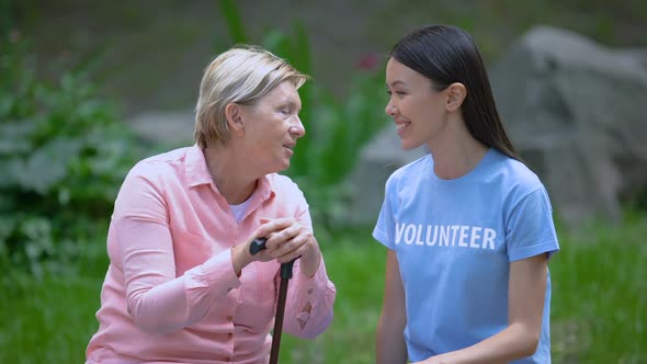 Senior Female Patient With Walking Stick Talking Young Volunteer Sitting Outdoor