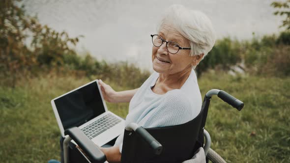 Happy Senior Woman in the Wheelchair Using Laptop and Smiling Towards Camera