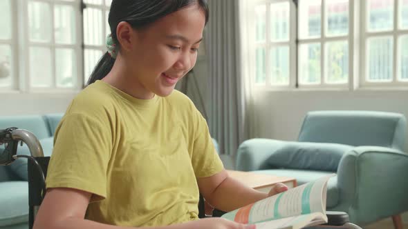 Close Up Of Asian Kid Girl Sitting In A Wheelchair And Reading Book At Home