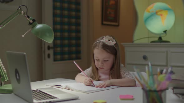 Cute Preschool Girl Writing Down in Notebook with Pen Looking at Laptop