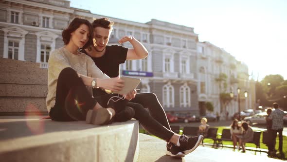 Young Beautiful Couple Smiling Speaking Looking at Tablet Sitting in City Park