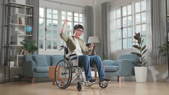 Asian Man Sitting In A Wheelchair While Use Mobile Phone Playing Video Game And Celebrating At Home