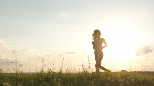 Young woman in light dress with long hair jogging on summer meadow at warm sunset.