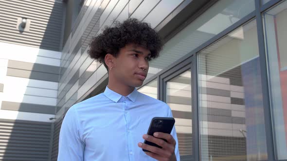 Guy in a Shirt Walks Through the Business District and Looks Into a Smartphone