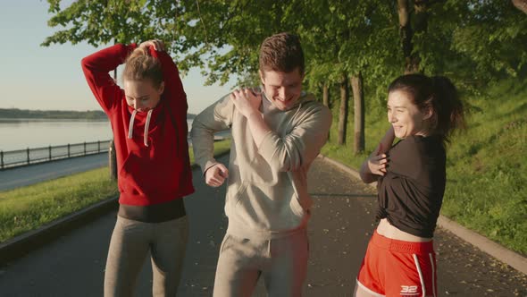 Doing Sport Together. Three Friends Stretching Muscles Before Jogging Along the Embankment of the