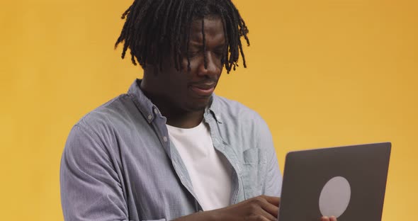 Studio Shot of Young African American Guy with Dreadlocks Browsing on Laptop  Orange Background