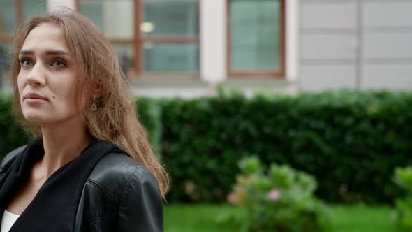 Portrait of a Blonde in a Black Jacket Against the Background of a Blurred City Street and Green