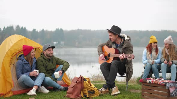 Group of Friends camping.They Are Sitting Around Camp Fire, Playing Guitar