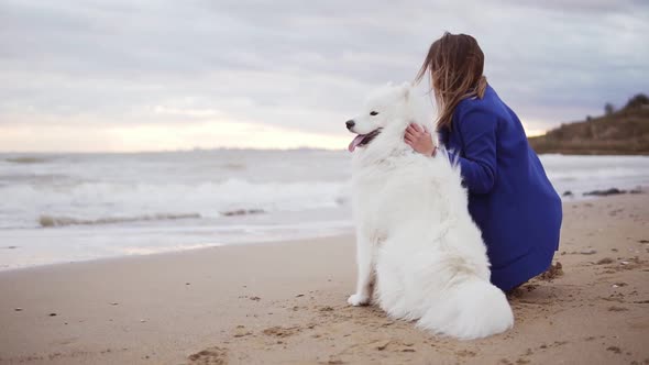 Back View of a Young Woman Sitting on the Sand and Embracing Her Dog of the Samoyed Breed By the Sea