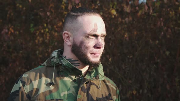 Soldier in Camouflage and Soot Strips on Face Looking on Sun Light and at Camera