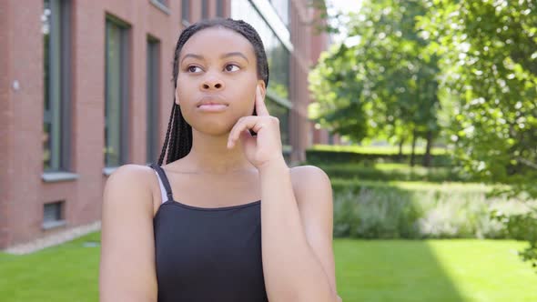 A Young Black Woman Thinks About Something - an Office Building and Green Park in the Background