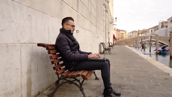 Young Man Works on Notebook Sitting on Old City Embankment