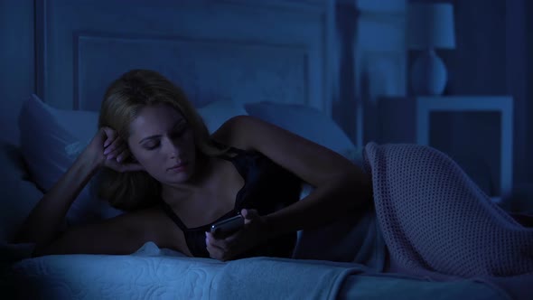 Beautiful Woman Lying in Bed, Reserving Disappointing Text Message on Smartphone