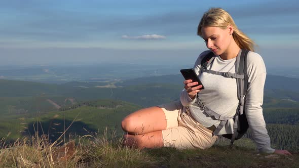 A Young Beautiful Woman Sits on Grass on a Hilltop and Works on a Smartphone