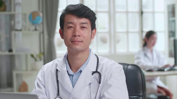 Young Asian Man Doctor Is Using Laptop Computer, He Turns And Smiles To Camera In Workplace