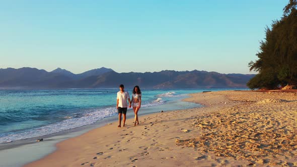 Happy couple walking barefoot on soft sand of exotic beach at sunrise with beautiful light over trop