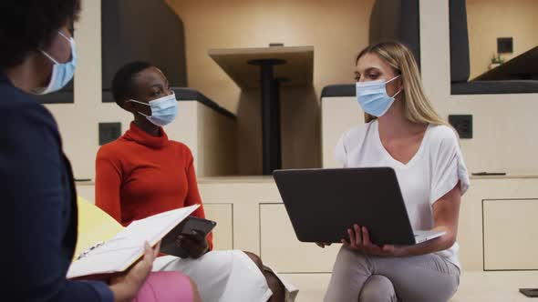 Three two diverse female office colleagues wearing face masks discussing together at office