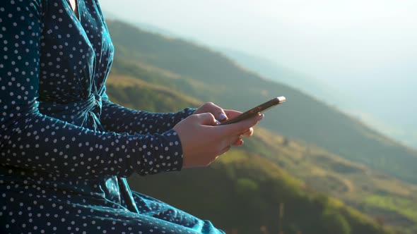 A Young Woman in a Dress Sits with a Phone in Her Hands Against a Background of Mountains Closeup