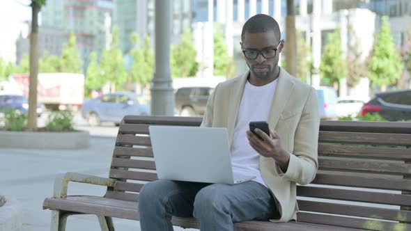 African Man Using Smartphone and Laptop While Sitting Outdoor on Bench