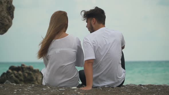 Back View Young Beautiful Couple Sits on Warm Pebbles on the Seashore