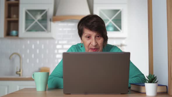 Happy Elderly Woman Communicating By Video Call Using Laptop at Home in Kitchen